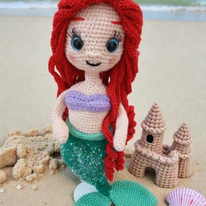 May include: A crocheted mermaid doll with red hair, a purple top, and a green sequined tail, posed on a sandy beach with a seashell and a crocheted sandcastle. The doll has large blue eyes and is smiling.