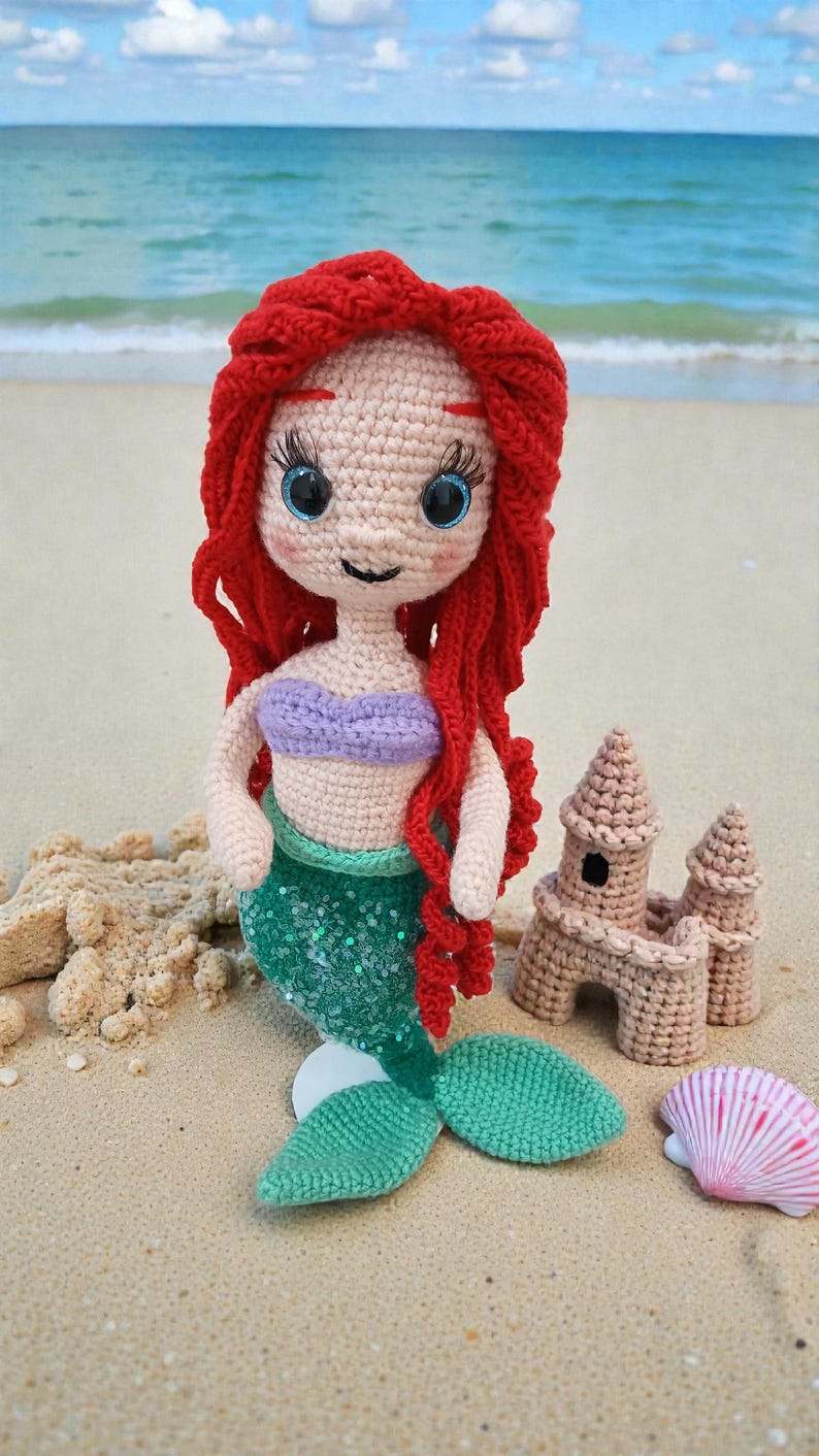May include: A crocheted mermaid doll with red hair, a purple top, and a green sequined tail, posed on a sandy beach with a seashell and a crocheted sandcastle. The doll has large blue eyes and is smiling.