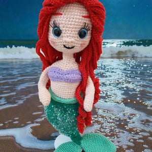 May include: Crocheted mermaid doll with red hair, blue eyes, and a purple top. The mermaid has a green sequined tail and is posed on a sandy beach with a moonlit ocean background. A handmade, whimsical item.