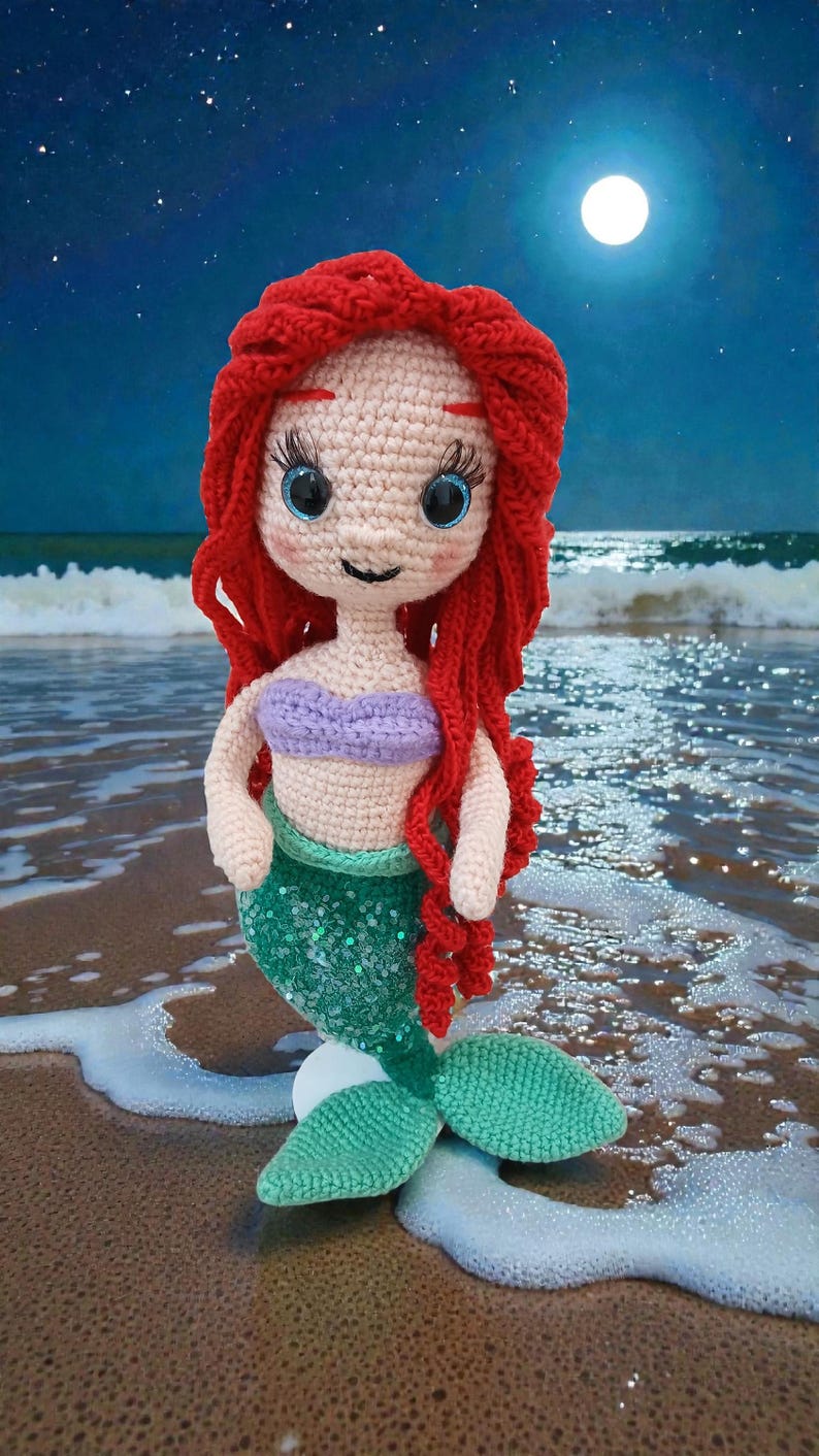May include: Crocheted mermaid doll with red hair, blue eyes, and a purple top. The mermaid has a green sequined tail and is posed on a sandy beach with a moonlit ocean background. A handmade, whimsical item.