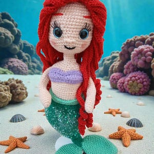 May include: A crocheted mermaid doll with red hair, blue eyes, and a purple top. The mermaid has a green sequined tail and is surrounded by seashells and starfish on a sandy surface. The background features an underwater scene with coral.