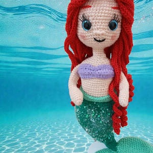 May include: Crocheted mermaid doll with red hair, blue eyes, and a purple top. The mermaid has a green sequined tail and is set against a blue water background, creating a whimsical underwater scene.
