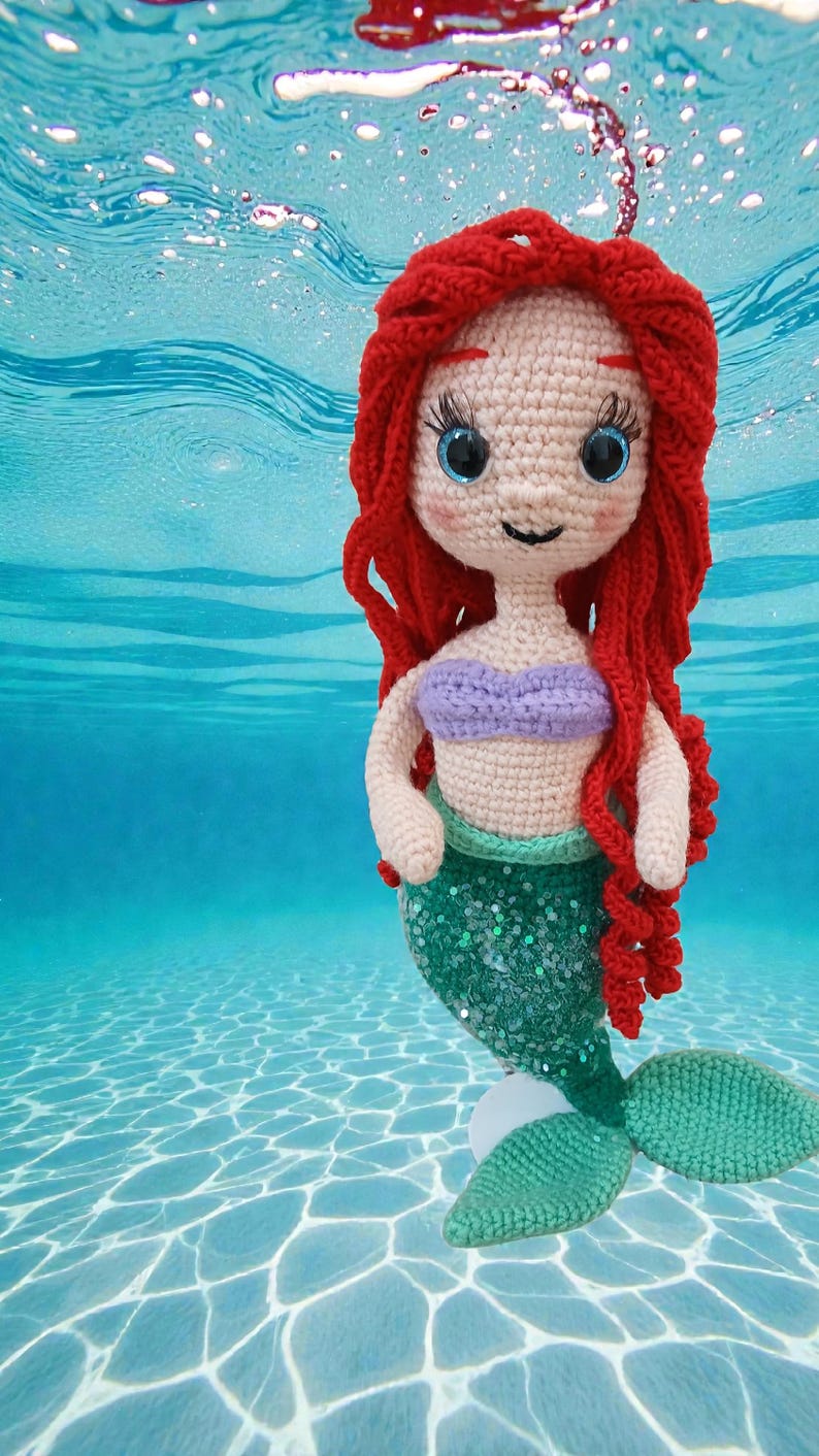 May include: Crocheted mermaid doll with red hair, blue eyes, and a purple top. The mermaid has a green sequined tail and is set against a blue water background, creating a whimsical underwater scene.