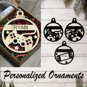 May include: Wooden Christmas ornament with a video game controller design, personalized with the name "RYAN" and the year "2024". The ornament is in a gift box with red and green filler. Additional black ornaments with game controller and handheld console designs.
