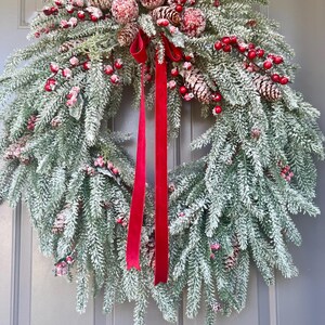 May include: A festive Christmas wreath with frosted green pine needles, red berries, and pinecones. The wreath is adorned with red ribbon and is displayed on a gray door. This holiday decoration adds a touch of seasonal cheer.