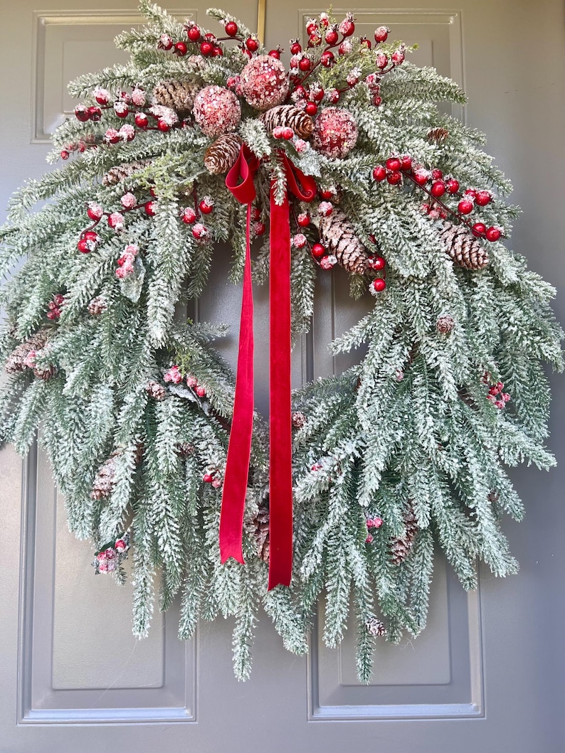 May include: A festive Christmas wreath with frosted green pine needles, red berries, and pinecones. The wreath is adorned with red ribbon and is displayed on a gray door. This holiday decoration adds a touch of seasonal cheer.
