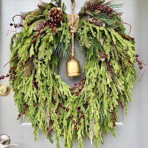 May include: A festive Christmas wreath with green pine needles, red berries, and pine cones. A small gold bell hangs from the center, suspended by a jute rope. The wreath is attached to a gray door.