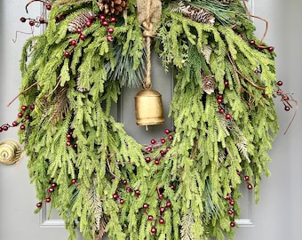 May include: A festive Christmas wreath with green pine needles, red berries, and pine cones. A small gold bell hangs from the center, suspended by a jute rope. The wreath is attached to a gray door.