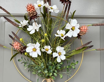 May include: A gold metal wreath with white flowers and brown pinecones. The wreath is tied with a brown twine bow.