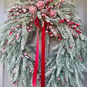May include: A festive Christmas wreath with frosted evergreen branches, red berries, pinecones, and red ribbon. The wreath is decorated with red and white ornaments, creating a winter holiday theme. The wreath is displayed on a gray door.