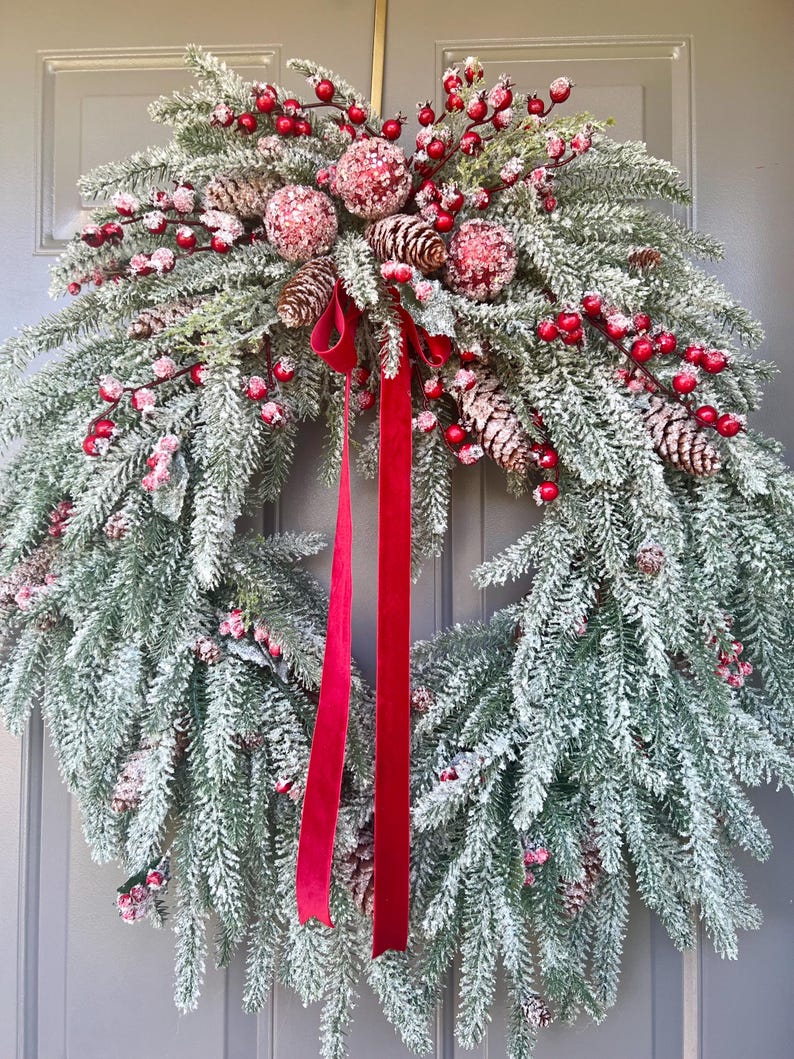 May include: A festive Christmas wreath with frosted evergreen branches, red berries, pinecones, and red ribbon. The wreath is decorated with red and white ornaments, creating a winter holiday theme. The wreath is displayed on a gray door.