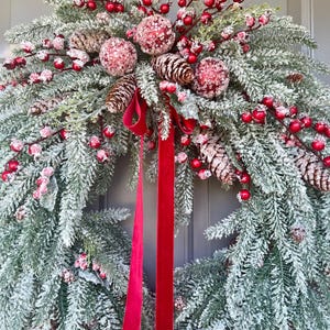 May include: A festive Christmas wreath with frosted evergreen branches, red berries, and pinecones. The wreath is adorned with three glittery red ornaments and a long, red velvet ribbon.