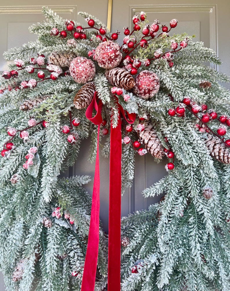 May include: A festive Christmas wreath with frosted evergreen branches, red berries, and pinecones. The wreath is adorned with three glittery red ornaments and a long, red velvet ribbon.