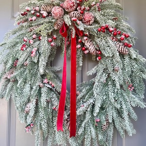 May include: A festive Christmas wreath with frosted evergreen branches, red berries, and pinecones. The wreath is adorned with red velvet ribbon, creating a classic holiday decoration. The wreath is displayed on a gray door.