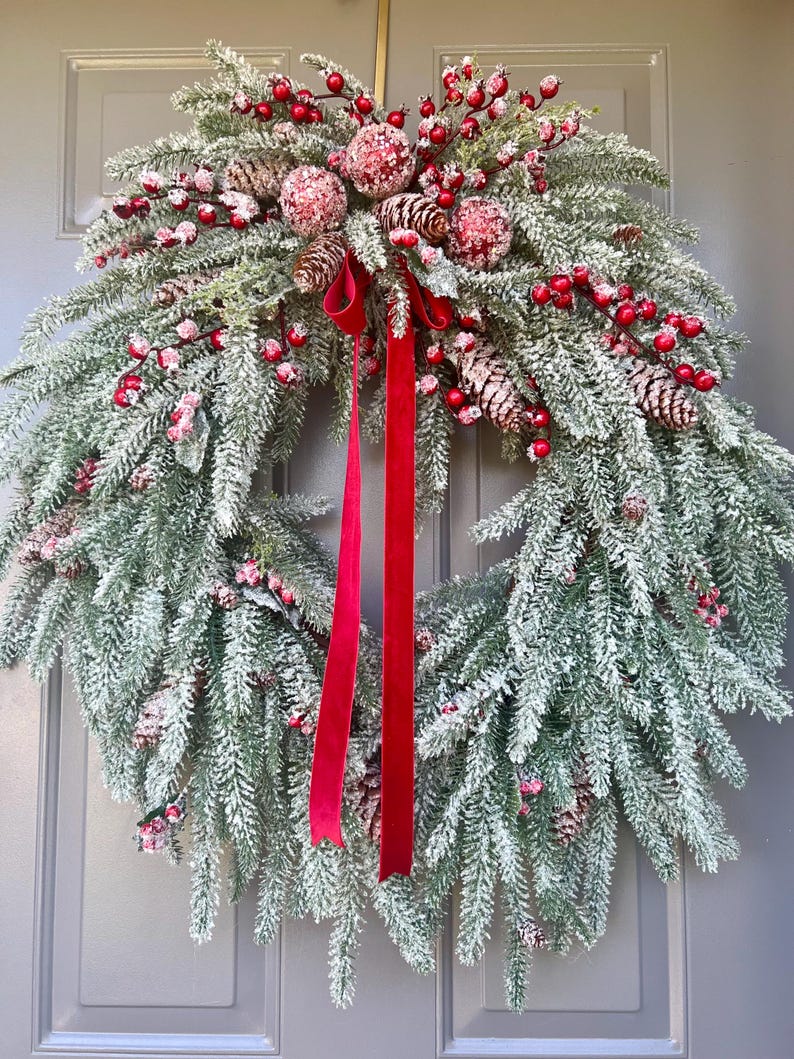 May include: A festive Christmas wreath with frosted evergreen branches, red berries, and pinecones. The wreath is adorned with red velvet ribbon, creating a classic holiday decoration. The wreath is displayed on a gray door.