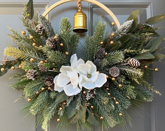 May include: A Christmas wreath made of pine branches, eucalyptus, and white magnolia flowers. The wreath is decorated with gold berries and pine cones and has a gold bell hanging from the top.