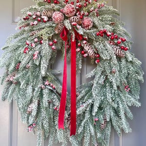 May include: A frosted evergreen Christmas wreath with red berries, pinecones, and decorative red spheres. A long, red velvet ribbon hangs from the wreath. The wreath is displayed on a gray door.