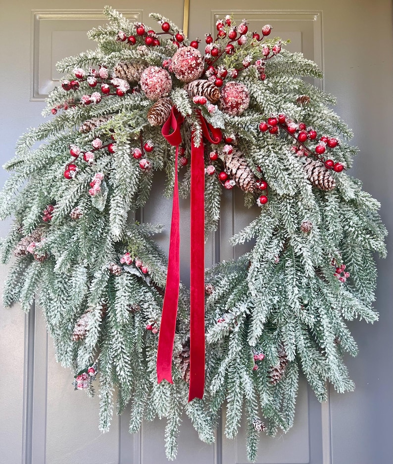 May include: A frosted evergreen Christmas wreath with red berries, pinecones, and decorative red spheres. A long, red velvet ribbon hangs from the wreath. The wreath is displayed on a gray door.
