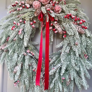 May include: A festive Christmas wreath with faux snow-covered evergreen branches, red berries, and pinecones. A bright red ribbon hangs from the wreath, adding a pop of color. This decorative wreath is perfect for holiday decor.