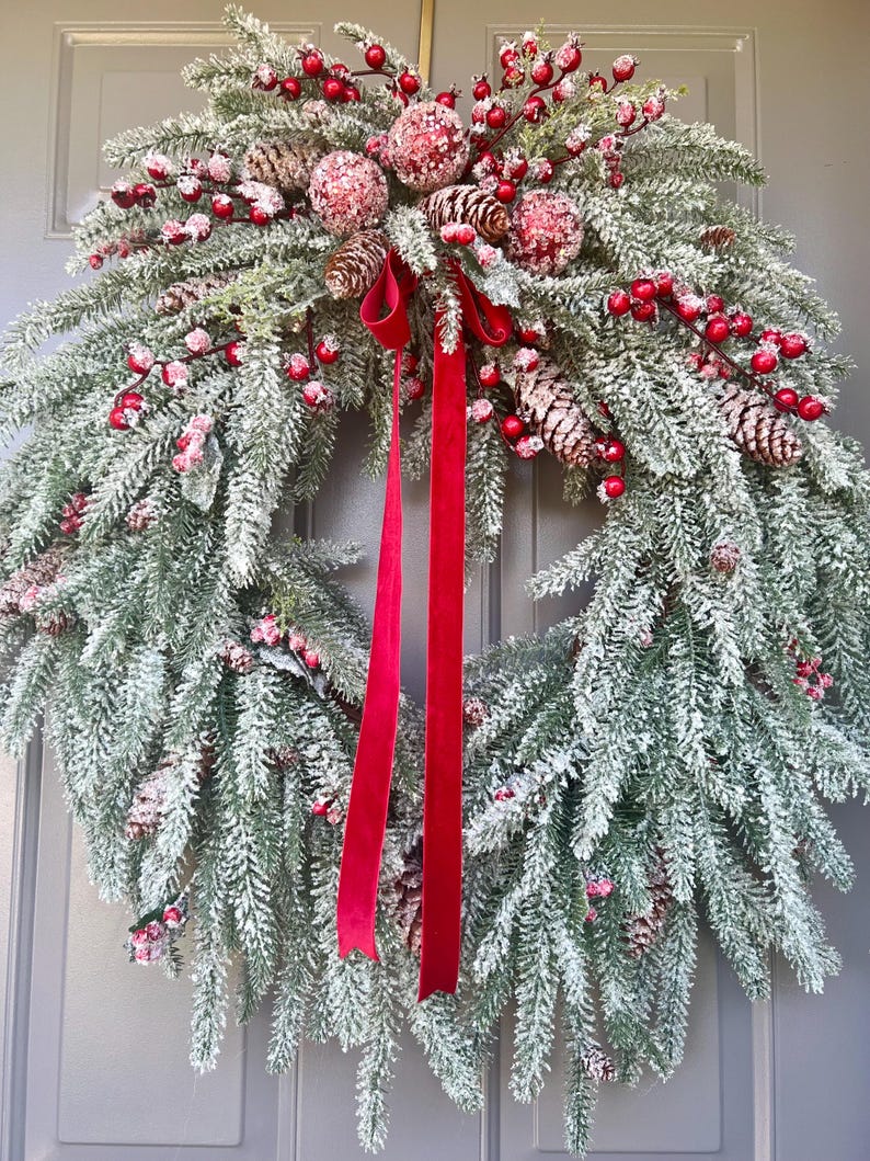 May include: A festive Christmas wreath with faux snow-covered evergreen branches, red berries, and pinecones. A bright red ribbon hangs from the wreath, adding a pop of color. This decorative wreath is perfect for holiday decor.