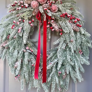 May include: A festive Christmas wreath with frosted evergreen branches, red berries, and pinecones. A bright red velvet ribbon hangs down the center. This holiday decoration is perfect for a front door or wall.
