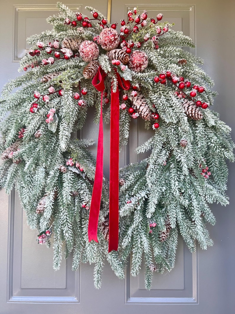 May include: A festive Christmas wreath with frosted evergreen branches, red berries, and pinecones. A bright red velvet ribbon hangs down the center. This holiday decoration is perfect for a front door or wall.