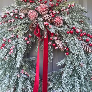May include: A festive Christmas wreath featuring frosted evergreen branches, red berries, and pinecones. The wreath is adorned with three glittery red ornaments and a long, red velvet ribbon. A seasonal decoration for the front door.