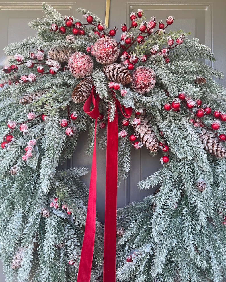 May include: A festive Christmas wreath featuring frosted evergreen branches, red berries, and pinecones. The wreath is adorned with three glittery red ornaments and a long, red velvet ribbon. A seasonal decoration for the front door.