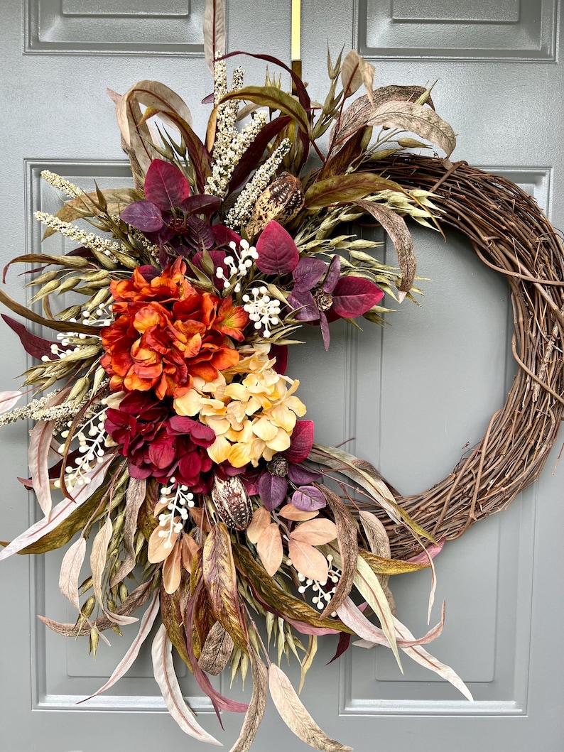 May include: Autumnal grapevine wreath featuring deep red, burnt orange, and beige hydrangeas, accented with dried seed pods, wheat stalks, and variegated eucalyptus leaves. The wreath is crafted with a rustic brown grapevine base, creating a fall-themed decorative piece suitable for front doors or walls.