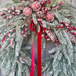 May include: A festive Christmas wreath featuring frosted evergreen branches, red berries, and pinecones. The wreath is adorned with three glittery pink ornaments and a vibrant red velvet ribbon. A seasonal decoration for the holiday season.