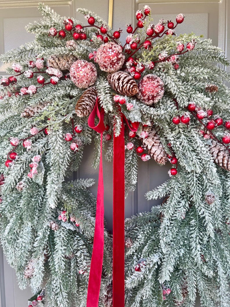 May include: A festive Christmas wreath featuring frosted evergreen branches, red berries, and pinecones. The wreath is adorned with three glittery pink ornaments and a vibrant red velvet ribbon. A seasonal decoration for the holiday season.
