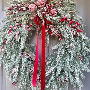 May include: A festive Christmas wreath with frosted green pine needles, red berries, and pinecones. The wreath is adorned with red velvet ribbon. This holiday decoration is perfect for adding a touch of seasonal cheer to any home.
