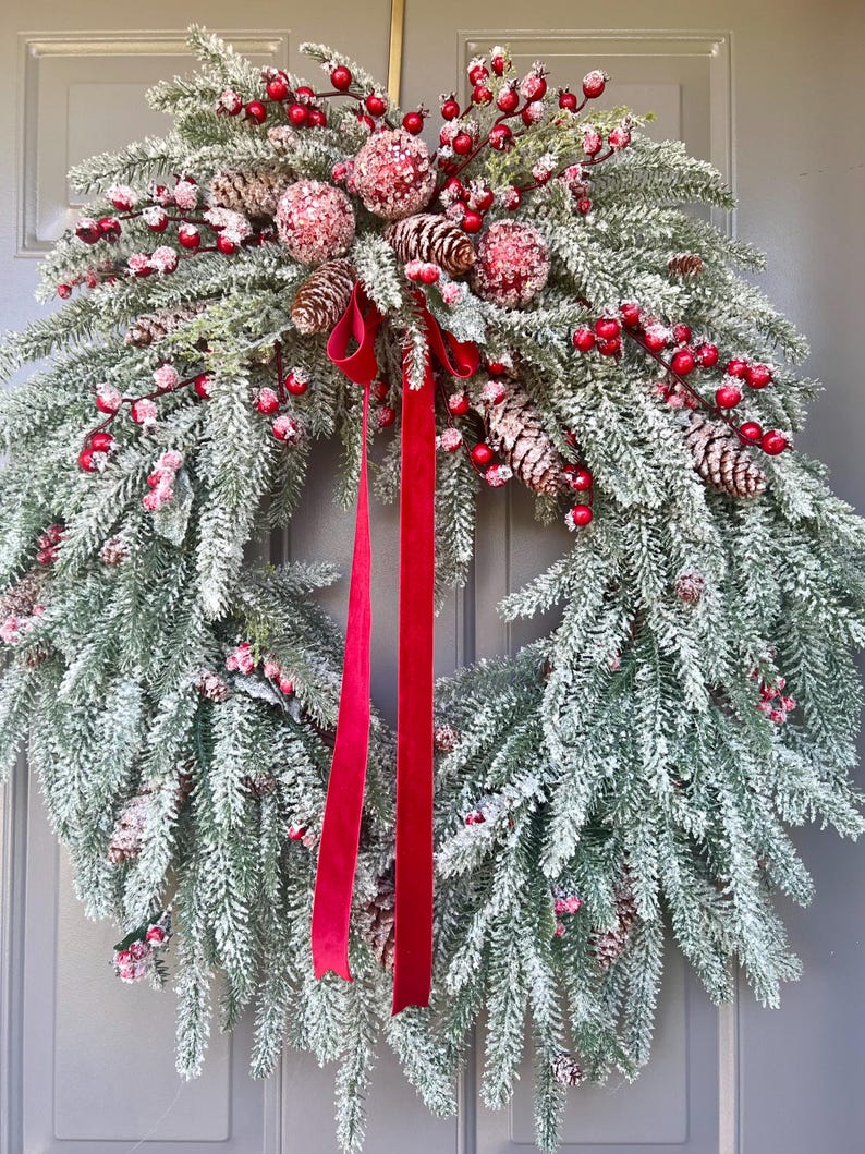 May include: A festive Christmas wreath with frosted green pine needles, red berries, and pinecones. The wreath is adorned with red velvet ribbon. This holiday decoration is perfect for adding a touch of seasonal cheer to any home.