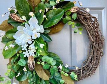 May include: A wreath made of grapevine branches with magnolia leaves, white flowers, and small white blossoms. The wreath is decorated with brown seed pods.