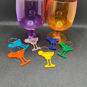 May include: Two stemware glasses, one purple and one orange, with matching colored stems. Six margarita-shaped wine charms in blue, orange, yellow, green, and clear are in front of the glasses. The glasses and charms are on a dark gray surface.
