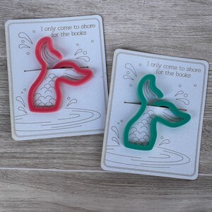 May include: Two mermaid tail shaped bookmark holders. One is pink and the other is green. Both have the text "I only come to shore for the books" on the card.