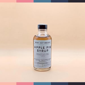 May include: A clear glass bottle of "Apple Pie Syrup" by Beef Cat Brews. The label lists ingredients like granulated sugar, maple, cinnamon, clove, allspice, and vanilla. The bottle has a black cap and is filled with a light brown liquid.