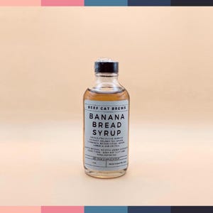 May include: A clear glass bottle of Banana Bread Syrup with a black cap. The label reads "BEEF CAT BREWS BANANA BREAD SYRUP". The syrup is light brown. The background is a light peach color.