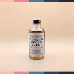 May include: A glass bottle of toasted pecan syrup with a black label that reads "Beef Cat Brews Toasted Pecan Syrup" and includes the ingredients: "Clarified Sugar, Oolong Tea, Toasted Pecan, Maple, Vanilla".