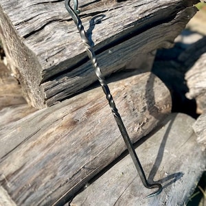 May include: A hand-forged, black metal fire poker with a twisted shaft and a hooked end. The poker is resting on weathered wooden logs, with the sunlight casting a shadow.