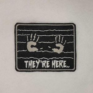 Könnte beinhalten: Schwarzer rechteckiger Aufnäher mit weißem Rand, der zwei graue Handabdrücke und den weißen Text "THEY'RE HERE.." zeigt. Das Design umfasst horizontale weiße Linien.