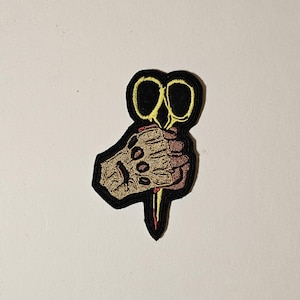May include: A black and yellow embroidered patch of a hand holding a pair of scissors with a bloody blade.