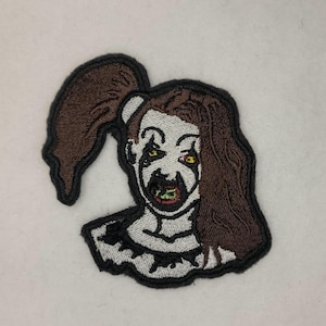 Può includere: Toppa ricamata di un clown inquietante con capelli castani in trecce, trucco bianco e nero e un sorriso sinistro.
