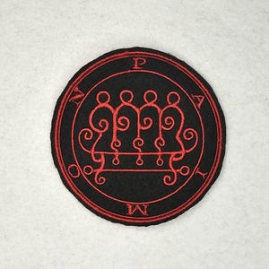 Può includere: Una toppa rotonda ricamata nera con un disegno di sigillo rosso. Il sigillo presenta un disegno centrale con tre figure collegate e testo circostante. La toppa è probabilmente per abbigliamento o accessori.