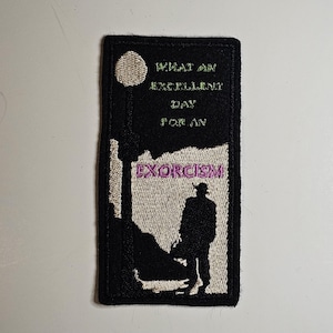 May include: Black and white embroidered patch with the text "What an excellent day for an exorcism". The patch features a silhouette of a person standing in front of a tree.