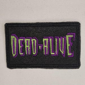 Puede incluir: Parche bordado negro con el texto "DEAD-ALIVE" en letras verdes y moradas.