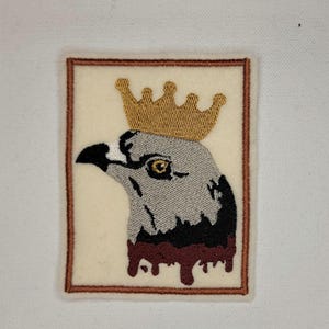 以下が含まれることがあります： 金色の冠をかぶった灰色のワシの刺繍パッチ。ワシの周りに茶色の塗料が垂れている。ワシは左を見ている。