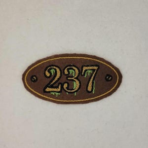 Puede incluir: Un parche ovalado marrón con un contorno amarillo y el número "237" bordado en amarillo con acentos verdes que gotean.