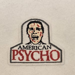 May include: A gray and red embroidered patch with a cartoon image of a man with blood on his face and the text "AMERICAN PSYCHO".