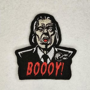 May include: Black and white embroidered patch of a man with a surprised expression, wearing a suit and tie. The text "BOOOOY!" is embroidered in red below the man.